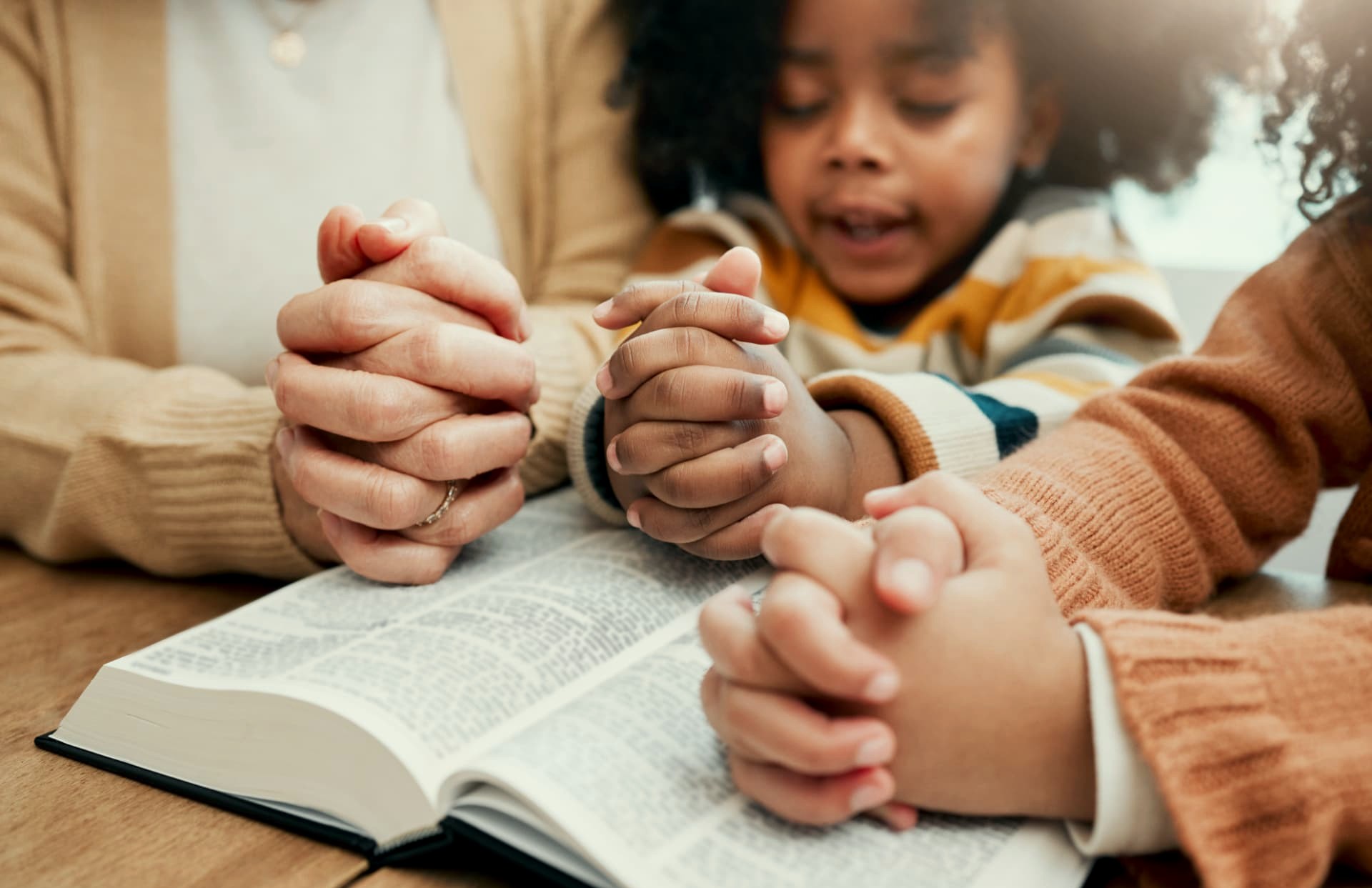 A parent and two children holding hands in prayer over an open Bible at home