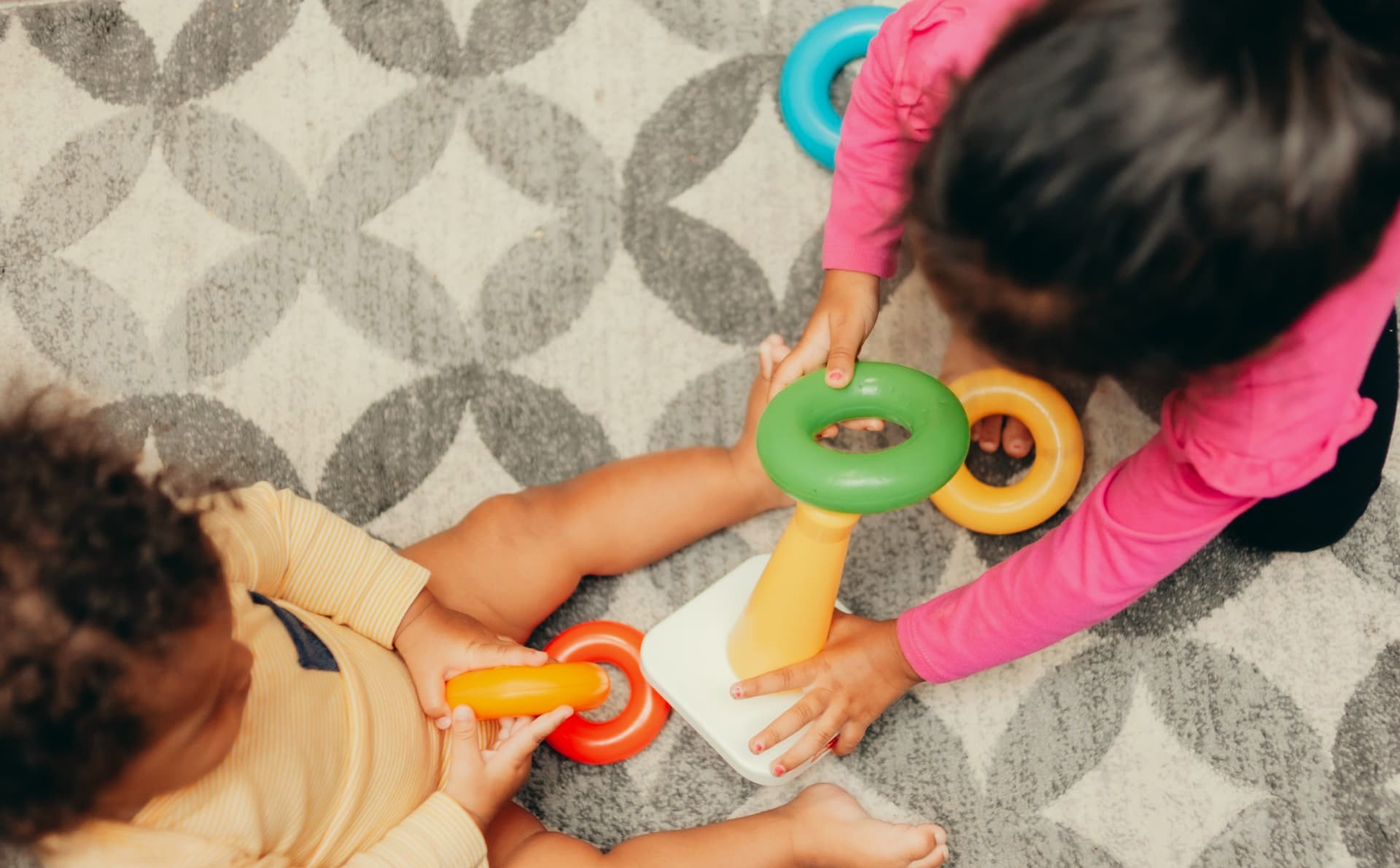 Two young children playing together with a ring-stacker toy on a soft rug